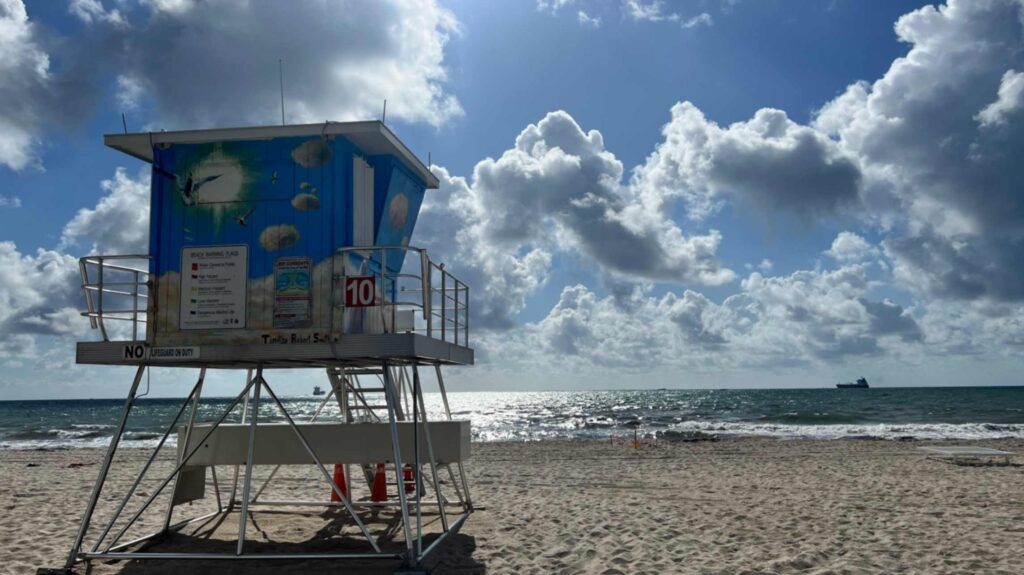 A painted blue lifeguard tower stands on a wide sandy beach under a dynamic sky filled with fluffy white clouds. The side of the tower features artwork of birds flying through a green sunburst and clouds, with a sign showing beach warning flags. The ocean water sparkles under the sunlight in the background where a cargo ship rests on the horizon.