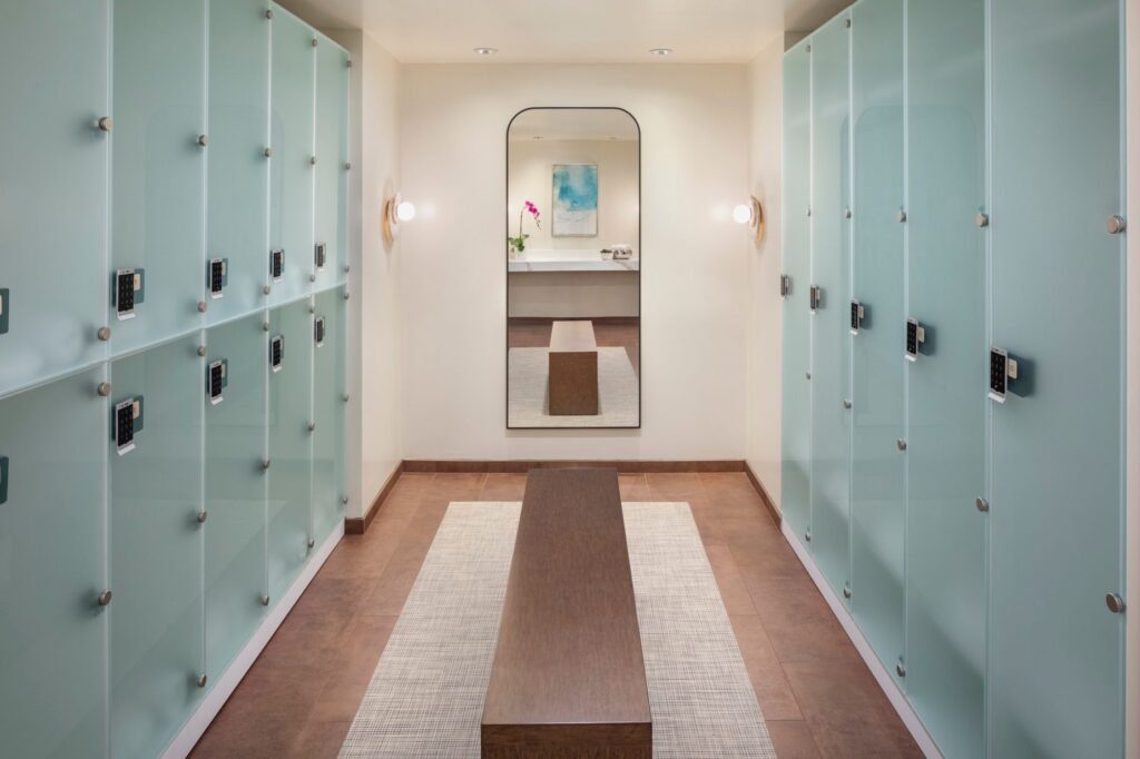 A modern spa locker room corridor lined on both sides with frosted glass lockers featuring digital keypads. A long wooden bench sits on a light woven rug over dark tiled floors in the center of the walkway. At the far end, a large arched mirror reflects a small vanity with an orchid and a piece of abstract blue wall art.