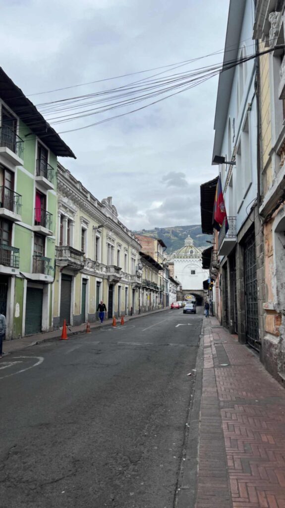 Street level view looking down a narrow colonial road in Quito lined with multi story historic buildings featuring iron balconies and the stone Arch of Santo Domingo framing a church dome in the far distance