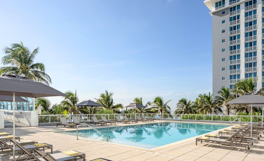 An outdoor resort swimming pool surrounded by a concrete patio deck with rows of grey lounge chairs and dark grey umbrellas. On the right, a modern multi-story hotel building rises into a clear blue sky, while palm trees frame the perimeter of the pool deck.