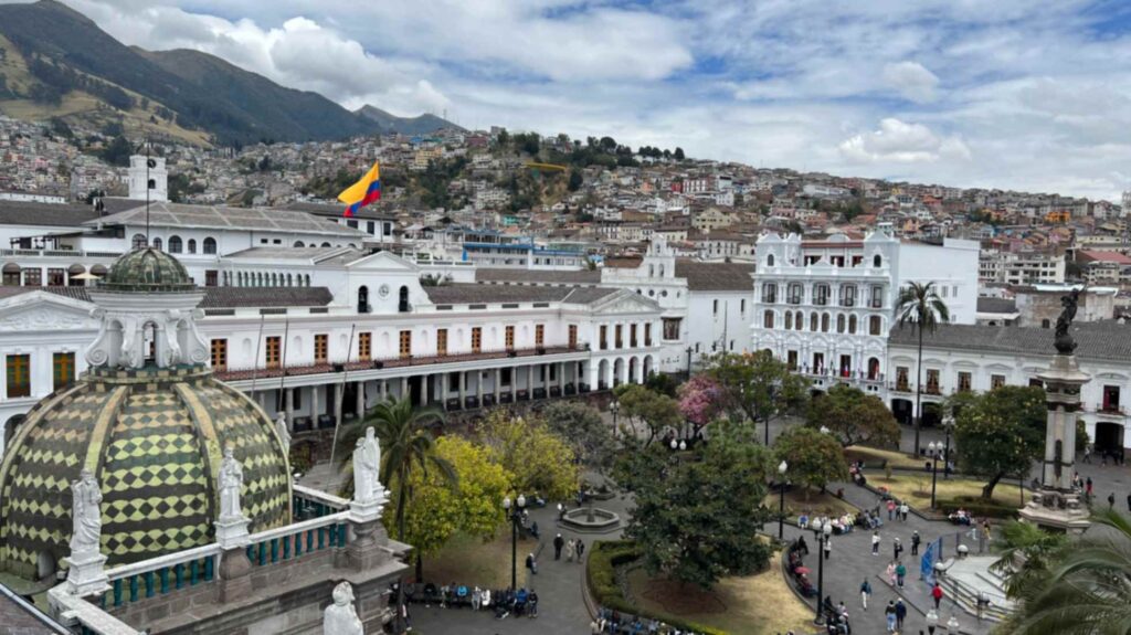 High angle view of Plaza de la Independencia in Quito Ecuador featuring historic white colonial buildings with arched walkways and a prominent green and yellow tiled dome in the foreground under a cloudy sky with hills rising in the background