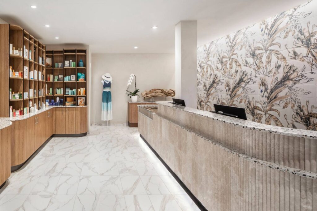 A bright and modern spa reception desk area featuring white marble tile flooring and a long textured beige counter. The wall behind the desk is decorated with a large wallpaper mural of neutral-toned tropical palm leaves. On the left, wooden shelves display various skincare and beauty products, with a blue and white sun dress displayed on a mannequin in the background.