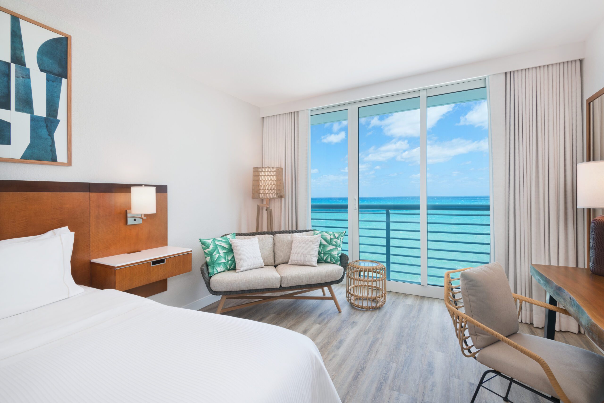 A bright hotel room corner featuring a small woven sofa with teal and white pillows next to floor-to-ceiling glass sliding doors. Outside the balcony railing, a wide expanse of turquoise and blue ocean meets a partly cloudy sky. The room includes light wood flooring, a wooden desk with a woven chair on the right, and the corner of a bed with white linens on the left.