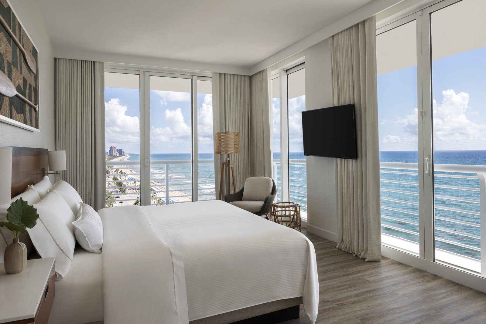 A spacious hotel guest room with multiple floor-to-ceiling glass doors showcasing wrap-around views of the sandy beach and ocean. The room features a king bed with crisp white bedding, an armchair nestled in the corner by a woven floor lamp, and a flat screen television mounted on the wall. Light grey curtains frame the bright windows under a partly cloudy sky.