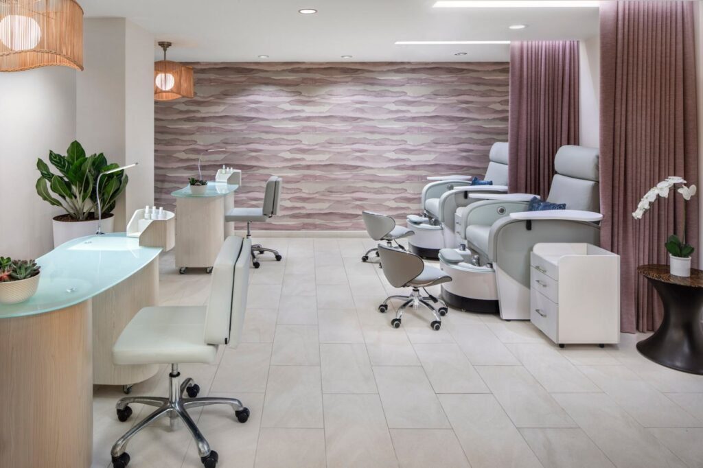 A chic hotel salon room featuring a wave patterned feature wall in muted pink and taupe tones. On the left, two glass top manicure stations are paired with white rolling chairs. On the right, plush grey pedicure chairs equipped with foot basins face rolling technician stools, framed by a soft mauve privacy curtain.