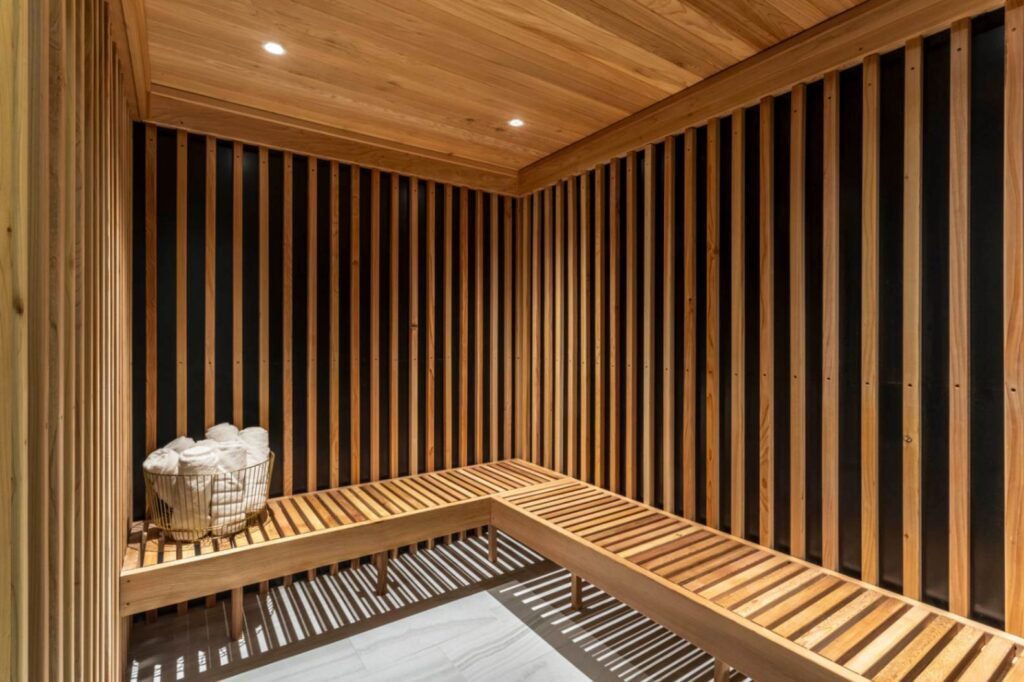 The interior of a modern wooden sauna featuring vertical light timber slats over dark backing walls. An L-shaped wooden bench runs along the walls above a light grey tiled floor, illuminated by soft recessed ceiling spotlights. A gold wire basket overflowing with rolled white towels rests on the left side of the bench.