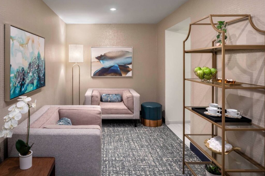 A serene spa relaxation lounge featuring warm beige walls, soft lighting, and two plush mauve armchairs arranged on a textured grey area rug. Large abstract paintings in coastal blue tones decorate the walls alongside a sleek metal floor lamp. On the right, a gold multi-tiered display shelf holds fresh green apples, teacups on a black tray, and rolled white towels.