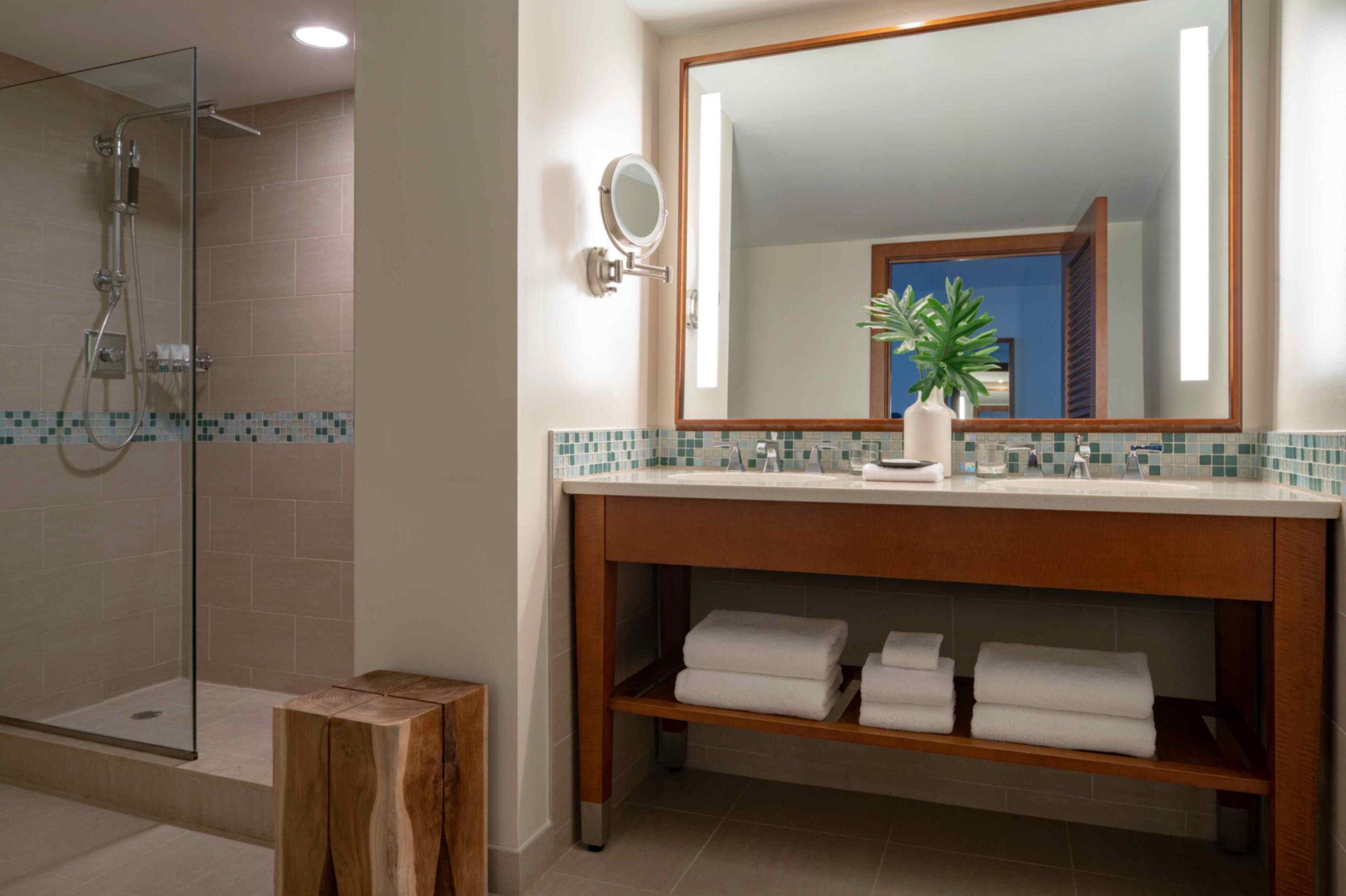 A modern hotel bathroom featuring a wooden double vanity with a light countertop and a large illuminated mirror. To the left, a walk in shower enclosed in glass displays beige tiles with a blue and green mosaic accent strip. Rolled white towels rest on the lower shelf of the vanity, and a rustic wooden block stool sits on the tiled floor.