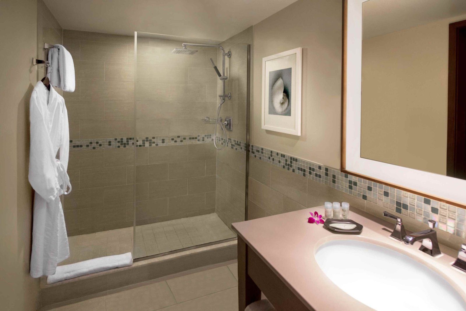 A hotel bathroom featuring a large glass enclosed walk in shower with beige wall tiles and a horizontal mosaic accent strip. A plush white bathrobe hangs on a wall hook next to the shower entrance. In the foreground, a vanity with a white undermount sink holds small toiletries and a purple flower blossom next to an illuminated mirror.