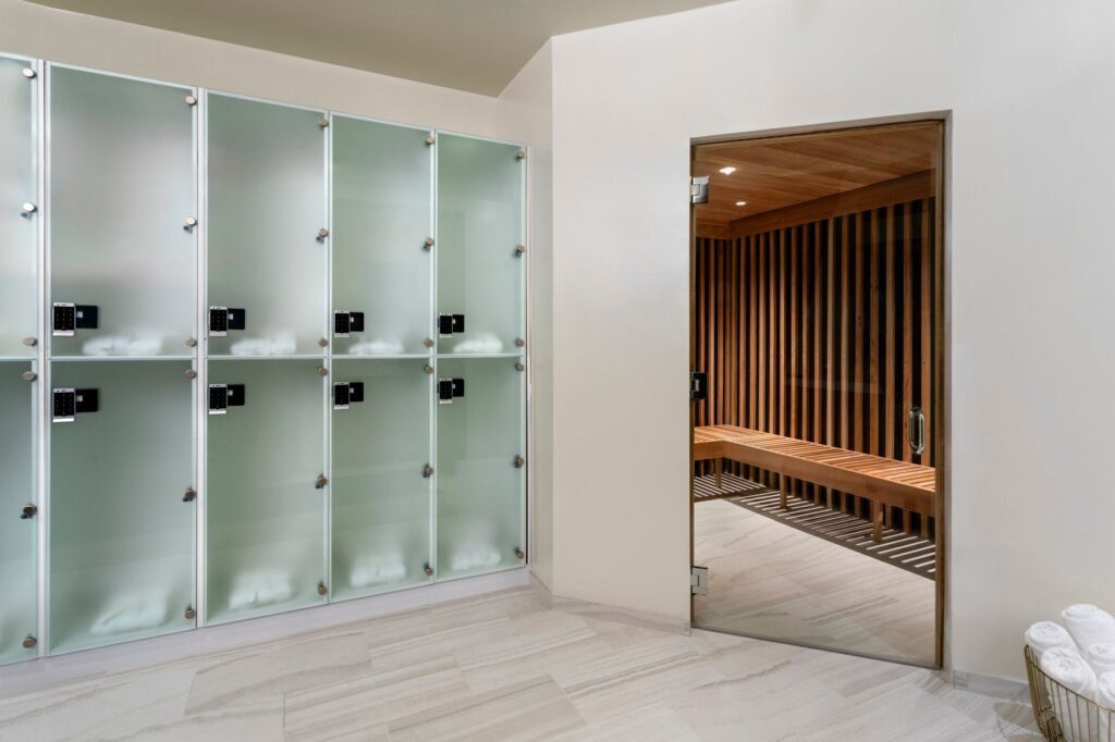 A bright spa locker room area with light marble tile flooring and frosted glass lockers featuring black digital locks. On the right, a glass door opens into a wooden sauna equipped with slatted timber walls and a long matching wood bench. A basket filled with rolled white towels rests in the lower right corner.