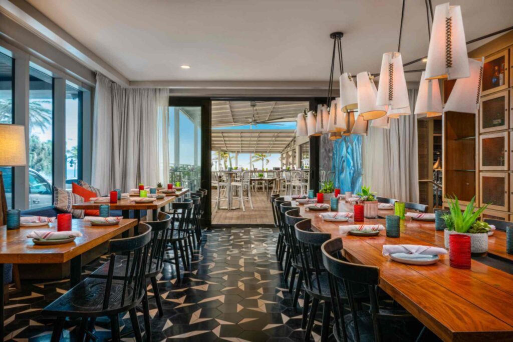 An interior view of a stylish resort restaurant dining room featuring a long wooden communal table and smaller perimeter tables set with vibrant glassware and striped napkins. The room boasts a bold black and white geometric tiled floor and clusters of white cone-shaped pendant lights, opening onto a sunlit outdoor patio deck with a glimpse of the ocean in the background.
