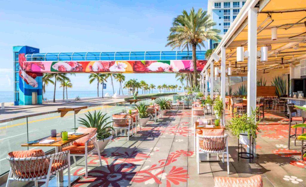 An open air restaurant patio overlooking a coastal road and the ocean. The patio features patterned floor tiles, modern dining tables, woven chairs with orange cushions, and potted plants. In the background, an elevated glass skywalk bridge decorated with vibrant red and pink floral art stretches toward the beach. Palm trees line the sandy shoreline under a bright blue sky.