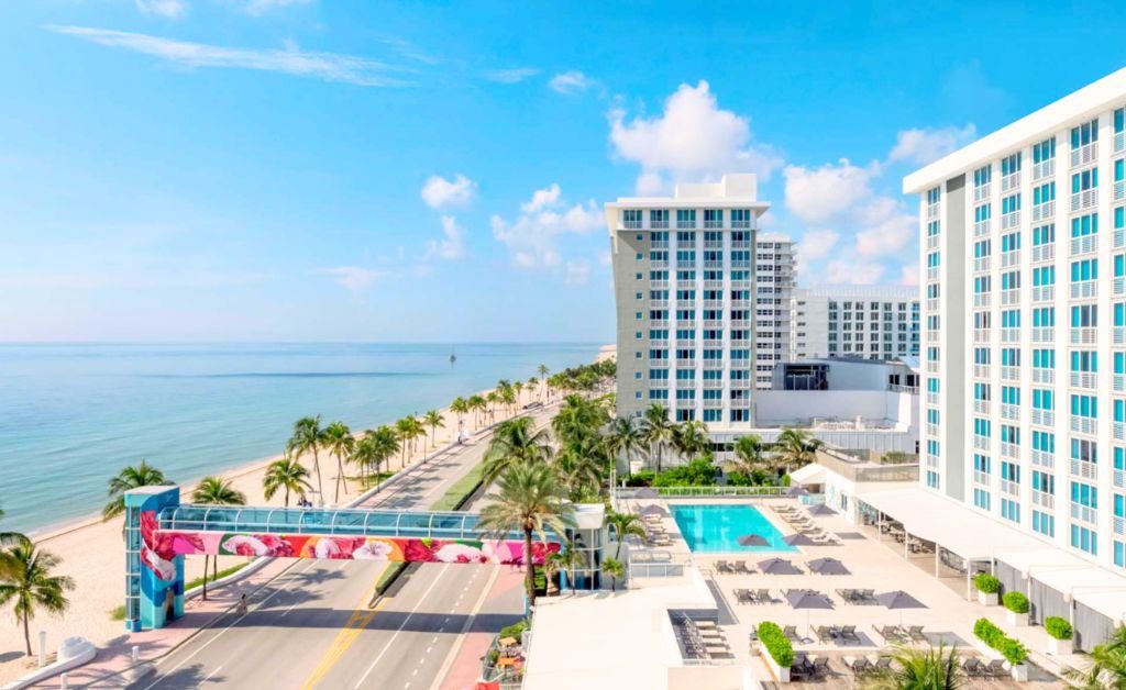 An elevated high angle view of a coastal resort pool deck and the adjacent beach. On the right, the multi story hotel building overlooks an outdoor swimming pool surrounded by gray lounge chairs and umbrellas. A coastal road runs between the resort and the sandy beach, spanned by an elevated blue skywalk bridge with vibrant floral art. Palm trees line the road and shoreline under a clear blue sky.